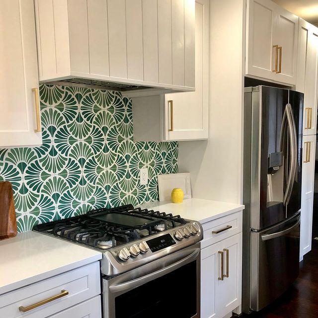 White kitchen cabinets kitchen, stainless steel appliances and a tiled flower themed backsplash in several shades of green. Dark wood floors.